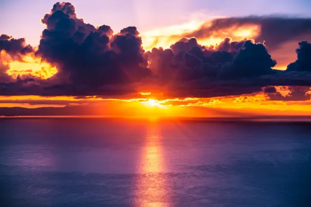 The sun setting over a calm sea, casting golden light through dramatic clouds.