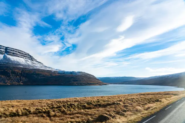 A blue fjord below a snow-capped mountain with golden grass and a road in the foreground, Iceland