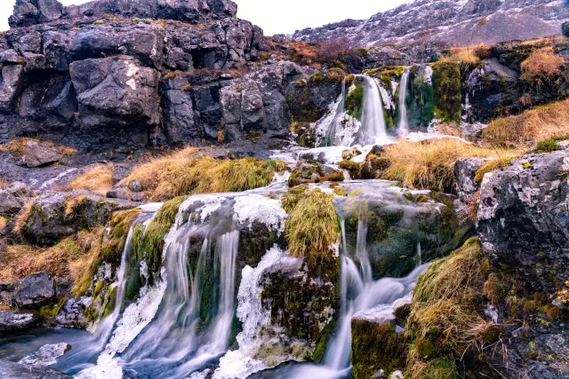 A small waterfall cascading over moss-covered rocks with ice formations in a rugged landscape.