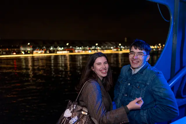 A couple posing by the water at night with city lights.